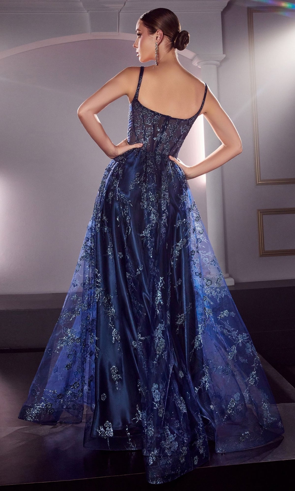A woman stands with her back to the camera, wearing the Long A-Line Glitter-Print Prom Dress J840 in deep blue with floral embroidery and a sheer overlay, her hair styled in a sleek bun.